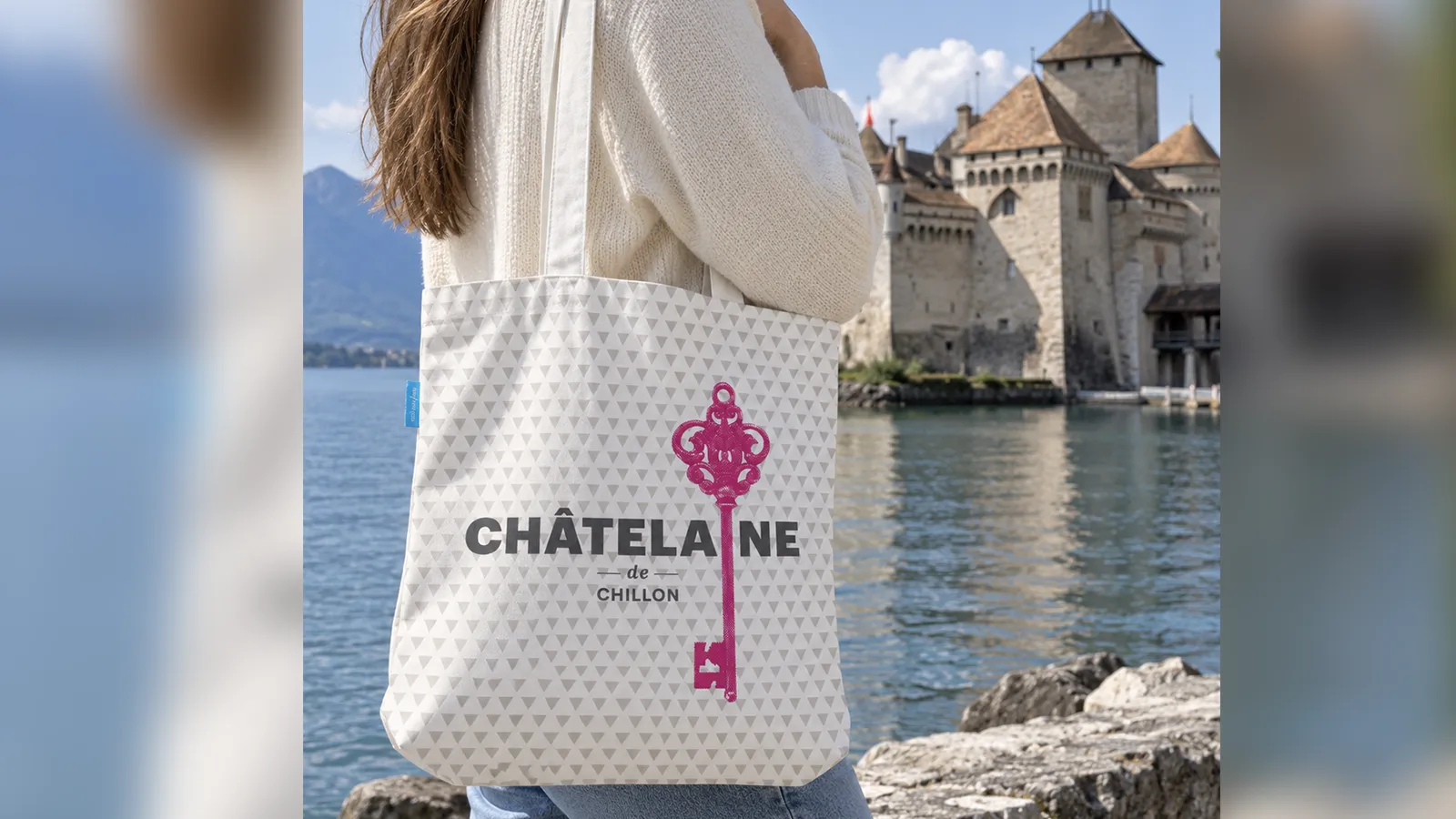 Femme portant le totebag « Châtelaine de Chillon », clé ancienne imprimée en rose sur un fond de petits triangles gris, devant le Château de Chillon et le lac Léman