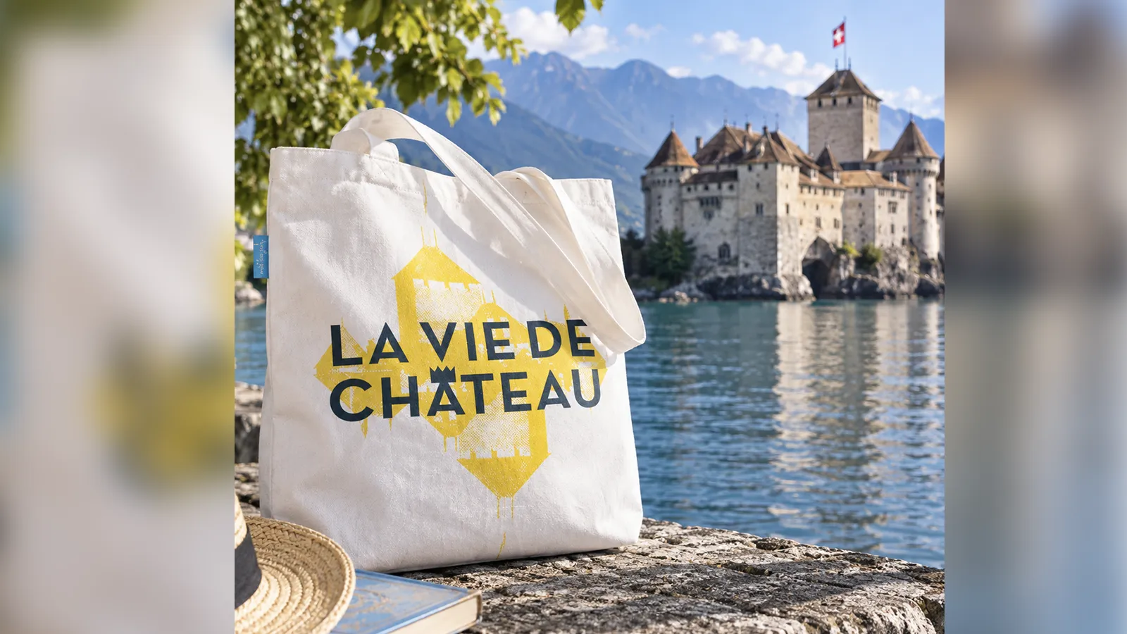 Totebag « La vie de château », lettrage bleu nuit sur silhouette jaune du château, posé sur un muret face au Château de Chillon et au lac Léman
