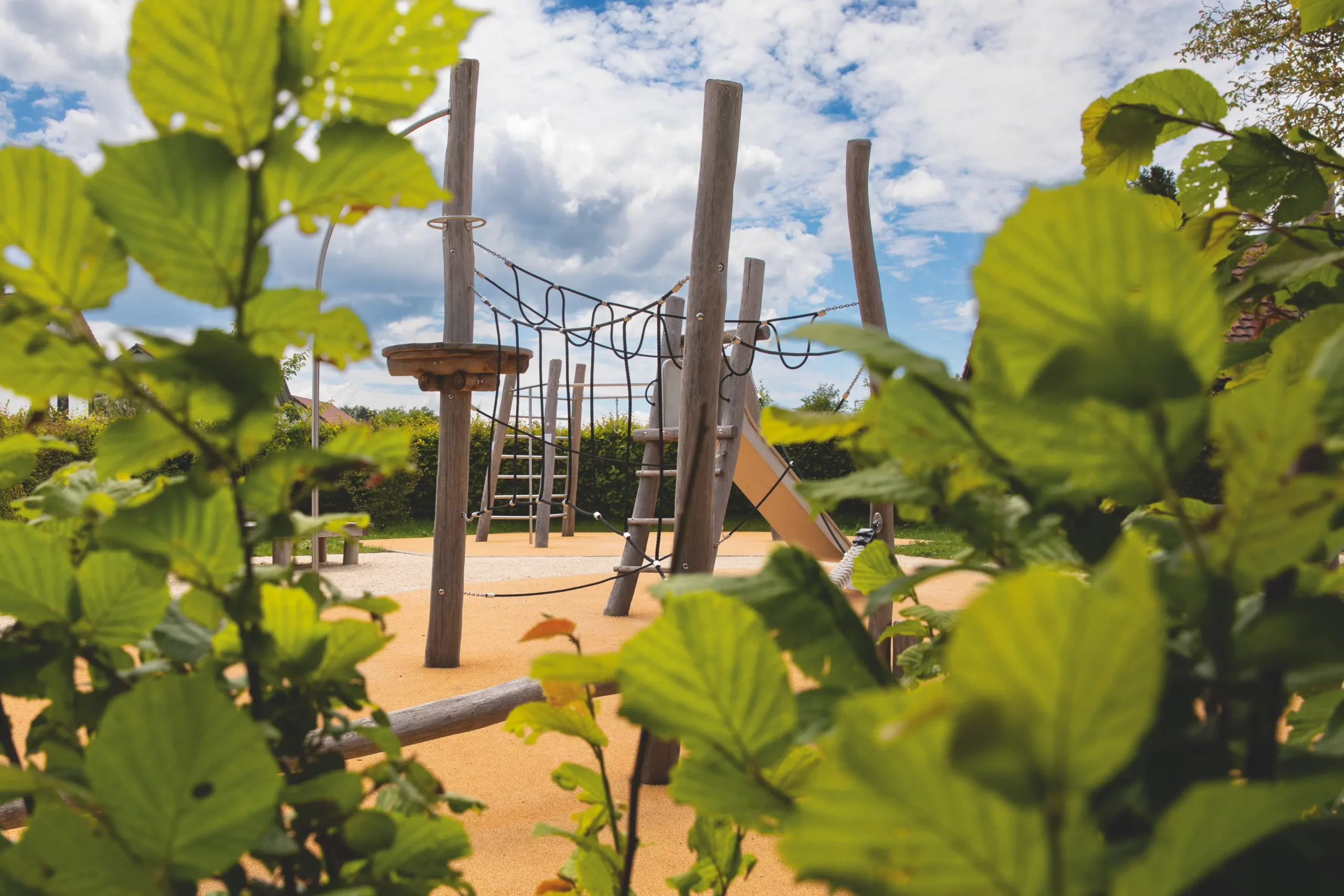 Aire de jeux en rondins de bois avec filets de corde, échelles et toboggan sur sol souple, entourée de haies, vue à travers un feuillage vert.